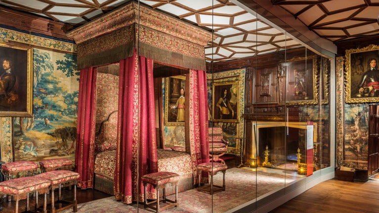 A room with a large framed ornate bed in the centre. The bed has red and gold covers and red curtains hanging at the corners. There is a fireplace to the right, stools to the left and at the foot of the bed and a tapestry on the wall to the left of the bed. There are also portraits hanging on the walls around the room, and the ceiling is white with a pattern created in wood.